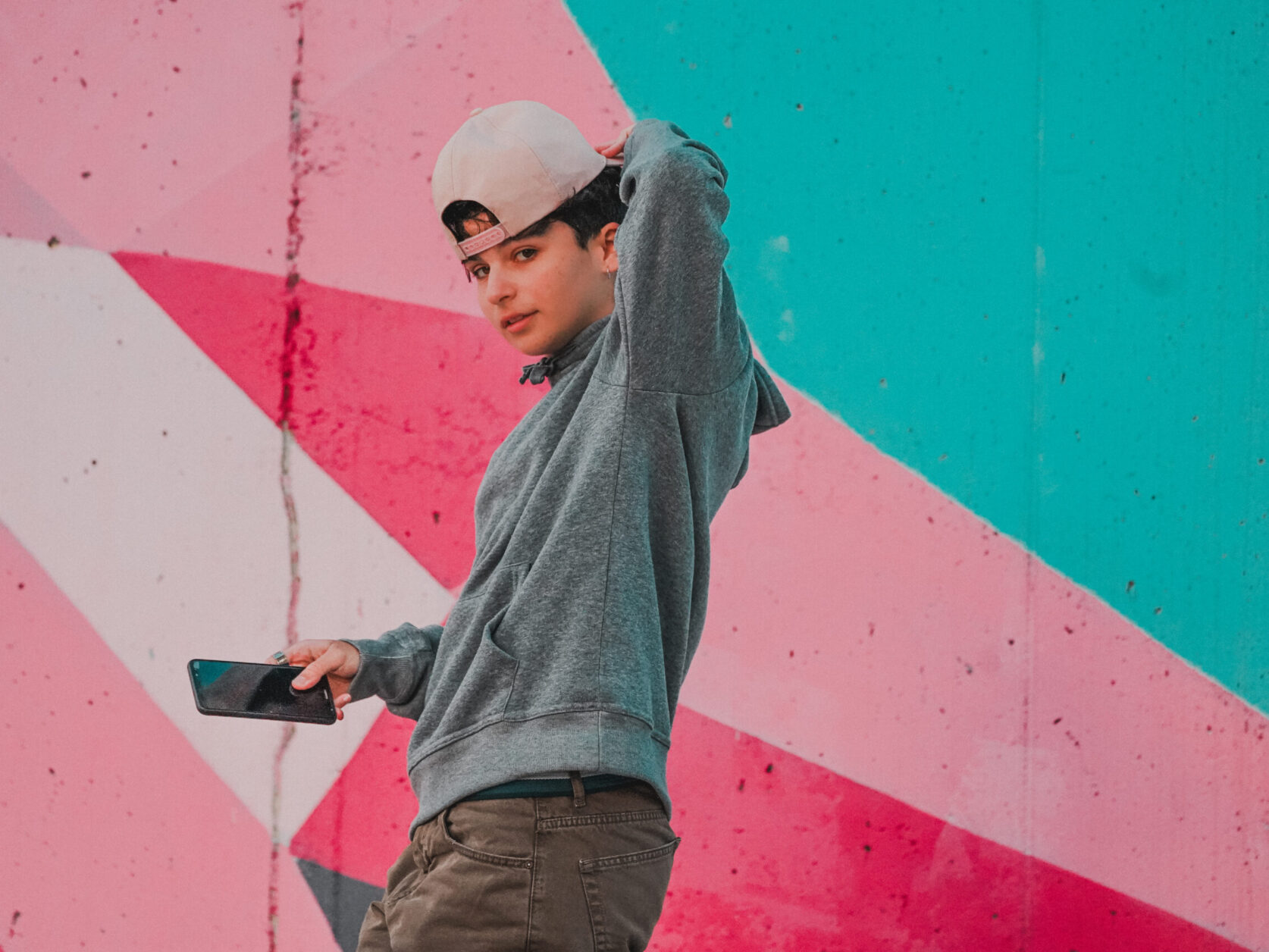 From below side view of trendy male teen in cap with raised arm and cellphone looking at camera near colorful wall